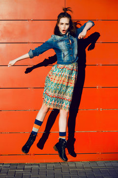 Young teenager girl having fun, posing and jumping near red wall background in skirt and jeans jacket on the sunset.