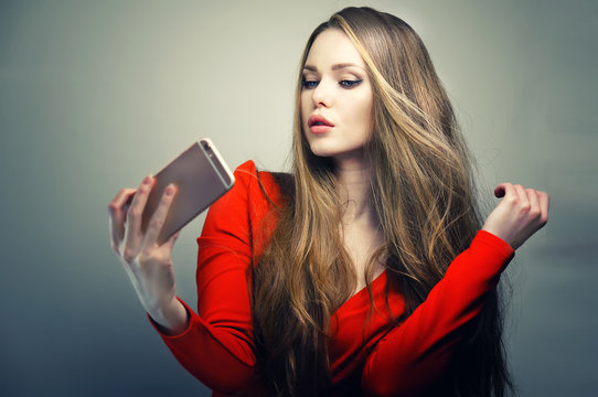 Beautiful woman in red dress with perfect cute face and blue eyes taking selfie with smartphone in studio.