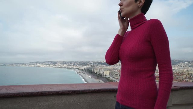 Female using app on the mobile phone. City view with sea and beach. Caucasian model dressed in elegant  jeans and red pullover. Sunny day in nice. Cote d'Azur. France. 