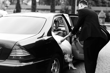 Groom helps bride to go out of the black Mercedes - obrazy, fototapety, plakaty