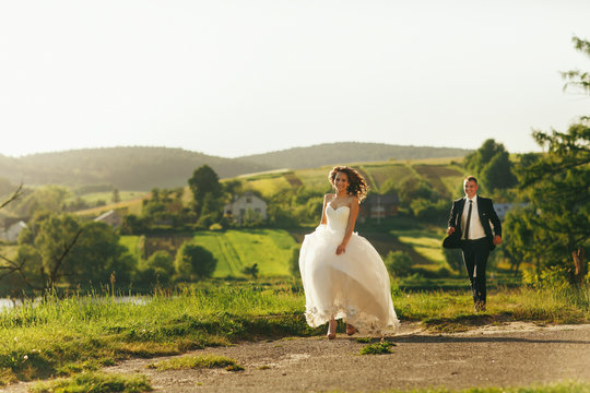 Bride And Groom Run Along The Village Road Enjoying Summer Weath