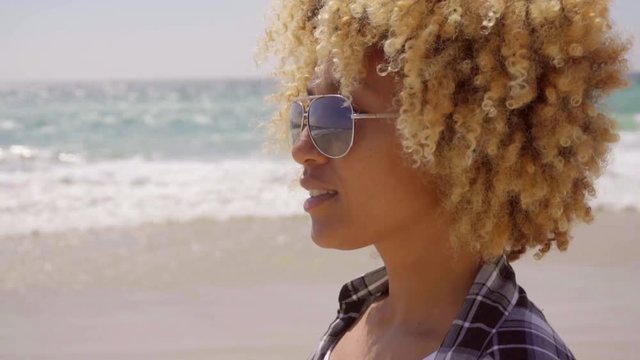 Attractive young woman in trendy sunglasses with a curly afro hairstyle walking on a deserted sandy beach in summer sunshine  close up cropped head and shoulders.