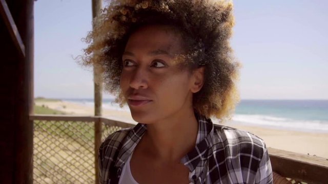 Adorable afro girl sitting on the terrace in kiters beach bar on windy day with marvelous beach view on her back