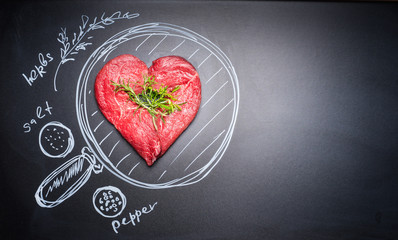Heart shaped chop of meat on black chalkboard with  painted pan and ingredients, top view, place...
