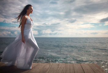 Fototapeta na wymiar Gorgeous woman in white dress standing at pier close to cliffs.