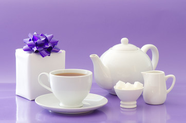 A white tea pot and cup of tea with a present on a lilac backgro