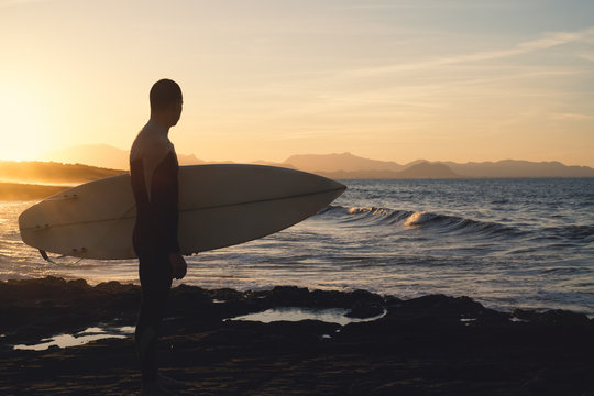 Young man surfer holding the surfboard while contemplating the waves at sunset.