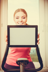 Woman on exercise bike holding pc tablet copyspace