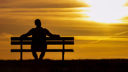 Silhouette man sitting on a bench looking up at the sunset  - 109902723