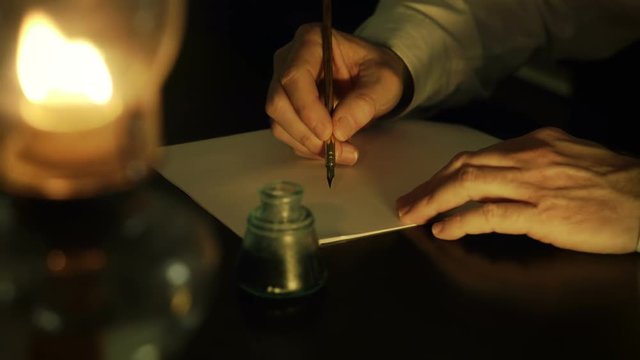 A man lit from the light of an oil lamp dips his pen in an inkwell and begins writing a letter. Closeup focus on hands.