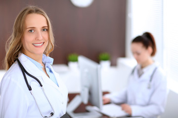 Beautiful young smiling female doctor standing in a hospital with her colleague in the background