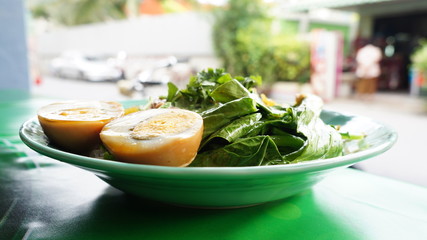 Green Vegetables and one halved boiled egg on a white plate in front of a street scene in Thailand.