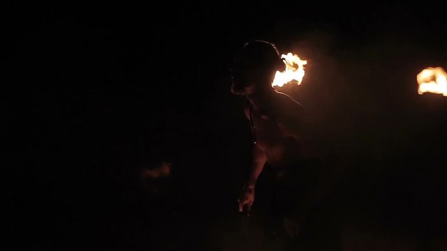 Fire show performance. Handsome male fire performer twirling fire baton and making fire breathing spitting flame against black background. Slow motion