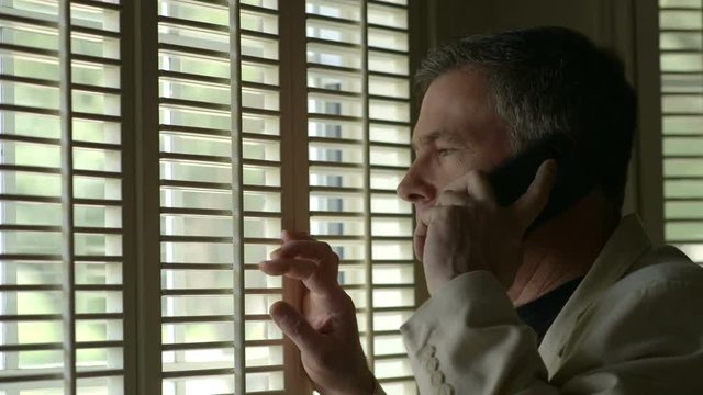 A man walks up to a shuttered window and looks out while he talks on his cell phone.