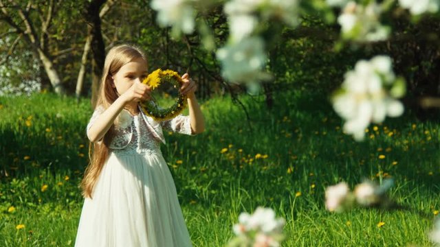 Portrait girl with yellow dandelions on the head spinning in the white dress in the park near blooming tree
