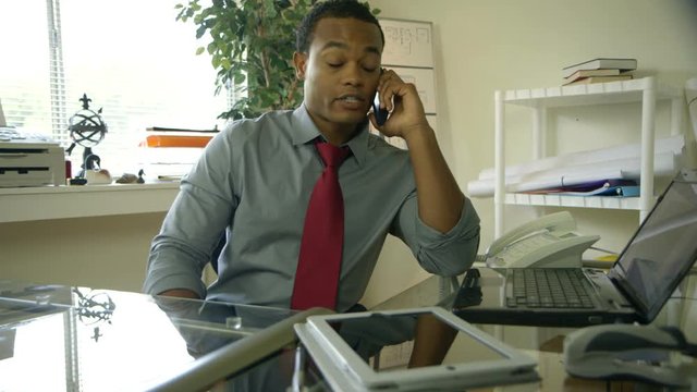 A young attractive African American man busy in his office gets a call on his cell phone and takes a file folder that is handed to him.