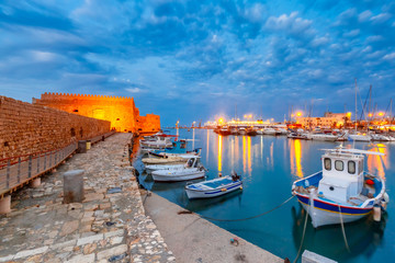 Old harbour of Heraklion with Venetian Koules Fortress, boats and marina during blue hour, Crete, Greece. Boats blurred motion on foreground. - obrazy, fototapety, plakaty