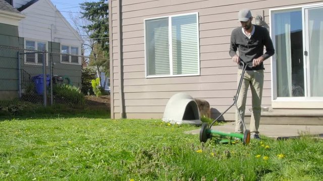 Attractive Mediterranean Man in baseball cap and beard mows a backyard lawn with a push mower 