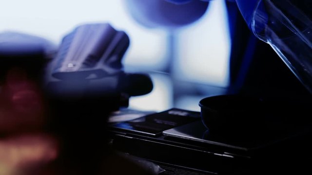 Sinister scene moves across laboratory paraphernalia to where a man with gun nearby is working with a crystallized substance.