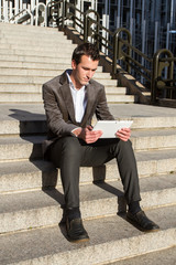 Businessman resting from work and while sending messages and talking with his white tablet. He is sitting on the stairs.