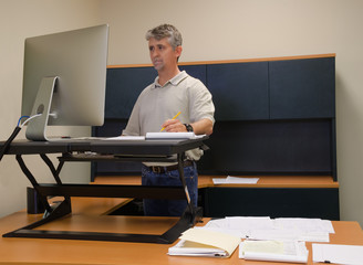 A man is working at a standup desk in an office where he works because standing is healthier than sitting all day. Live healthy, don’t sit all day. - 109968754