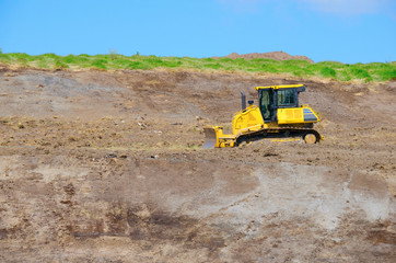A yellow bulldozer tractor is plowing on the side of a steep dirt hill with grass on top and blue sky in background.