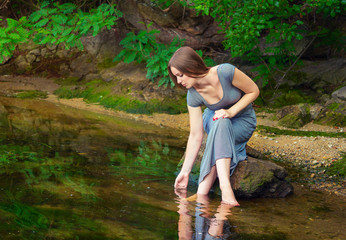 Naklejka na ściany i meble Beautiful young woman with long hair wearing casual dress sitting on the rock with feet in a weedy pond water