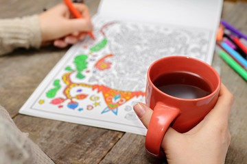 Fototapeta na wymiar Woman coloring an adult coloring book and drinking tea, new stress reliving tool