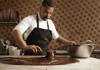 Black man professional chief collects tasty melted chocolate from vintage marble table to big steel pot in his artisan rustic kitchen with industrial retro machines. Mirrored in chocolate on table. - obrazy, fototapety, plakaty