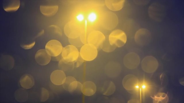 Traveling de farolas de iluminacion en carretera, tras la luna delantera del coche en movimiento ,con reflejos en las gotas de agua de lluvia en tono amarillento, al anochecer
