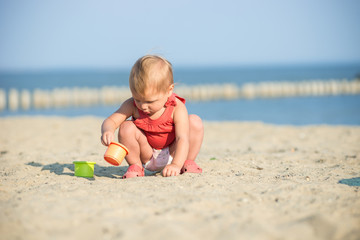 Baby playing on the sandy beach near the sea. Cute little girl in red dress with sand on tropical beach. Ocean coast.
