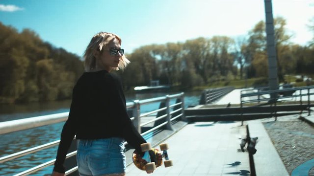 Young attractive Caucasian blonde girl walking with a skateboard in the city. 60 FPS slow motion shot. Blackmagic URSA Mini graded footage