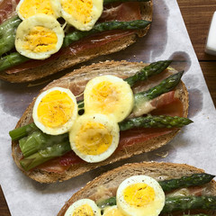 Gebackenes Vollkornbrot belegt mit Schinken, grünem Spargel, hartgekochtes Ei und geriebenem Käse, fotografiert mit natürlichem Licht