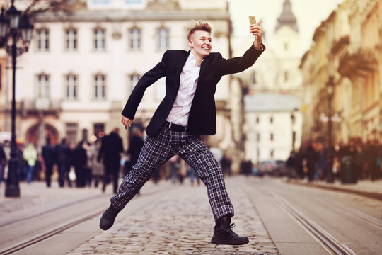 Full body portrait of young happy smiling handsome man running, jumping & taking selfie photo on old street. City lifestyle. Old european architecture on background. Toned style instagram filters
