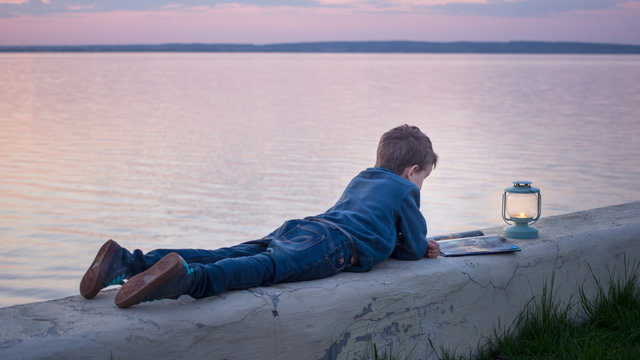 Child lying & reading a book at sunset near water