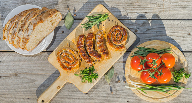Assorted grilled sausages served on wooden plate over wooden table with vegetables and bread on background. Overhead shot. Banner.