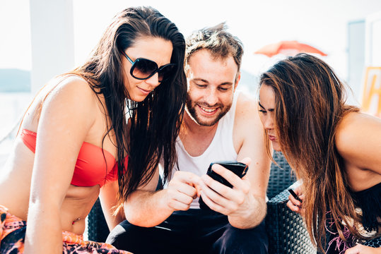 Group of young beautiful multiethnic woman and man friends sitting in a bar on the beach using smart phone handhold, chatting - technology, social network, communication concept
