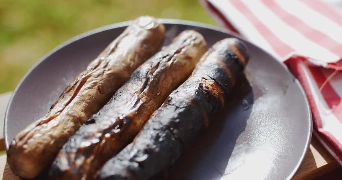 Row of three charred sausages on wooden board outside next to napkin with grass in the background