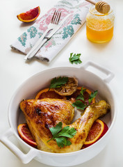Roasted chicken legs, red oranges, garlic and parsley in white baking dish on white table. Cooked with sauce from orange, honey and olive oil.