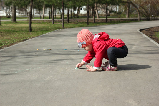 2-year-old girl in a red jacket draws with chalk on the pavement
