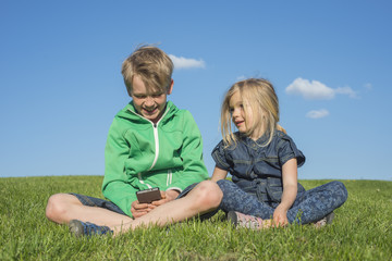 Naklejka na ściany i meble Happy blond children using smartphone (watching movie or playing game) sitting on the grass. Brother and sister. Summertime