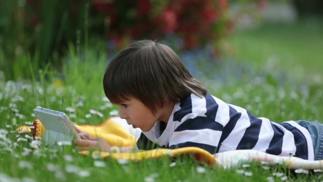 Sweet little boy, lying down in spring flower garden on a colorful blanket, playing on tablet