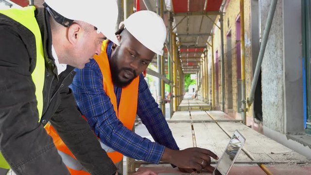 Side view: two architects, a black and a white, wearing safety jackets, helmets and goggles discussing looking at the computer among scaffolding
