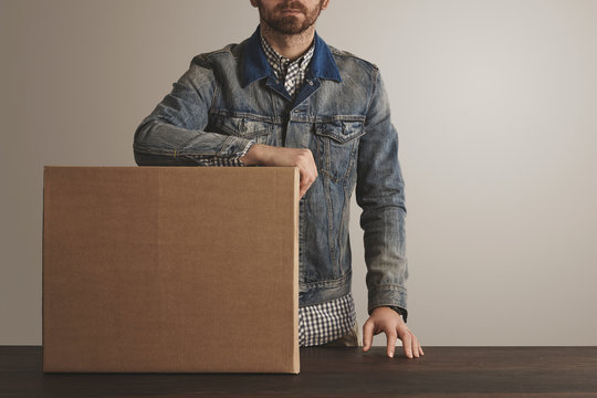 Bearded brutal courier in jeans work jacket stays near presented big carton paper box with goods on wooden table. Special delivery, retail shipping post box