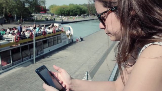 Girl Using Smartphone In Seine River With Boat Tour In Background, Paris. Woman using a smartphone on a sunny day in the famous Seine river with tourists, France