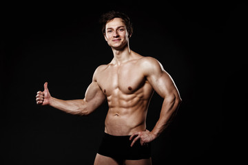 Fototapeta na wymiar Muscular and fit young bodybuilder fitness male model showing thumb-up and posing over black background. Perfect fitness body. Ideal for commercial.