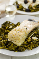codfish with olives and greens on white plate on white wooden background
