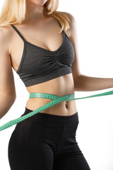 Closeup portrait of attractive cheerful fitness girl measuring her waist with tape in gray top and black leggings with red dumbbells posing over white background isolated 