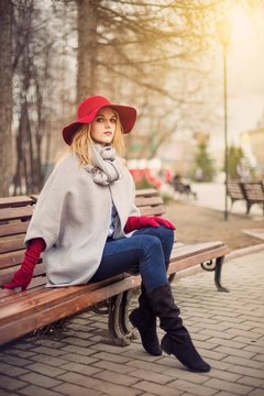 Portrait of young female in elegant red hat sitting on bench in city park