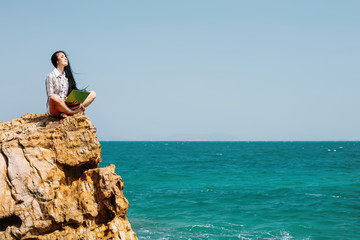 Fototapeta na wymiar Beautiful business woman sitting on stone cliff on blue sea beach, enjoy freedom, using laptop computer
